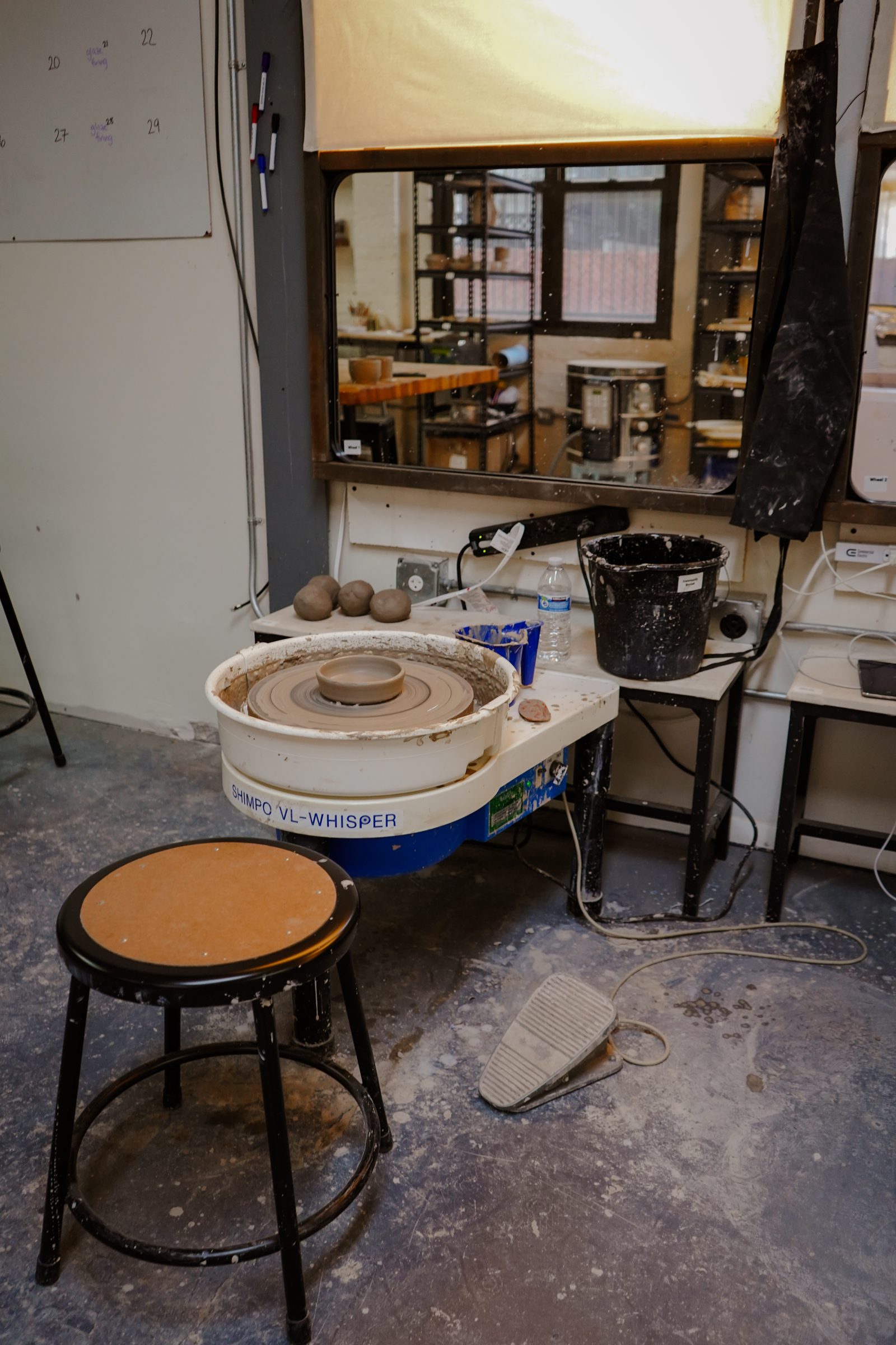 A Shimpo Whisper wheel and stool at a work station, with the firing schedule board behind